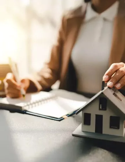 Conseillère immobilière tenant une maison miniature et prenant des notes à son bureau